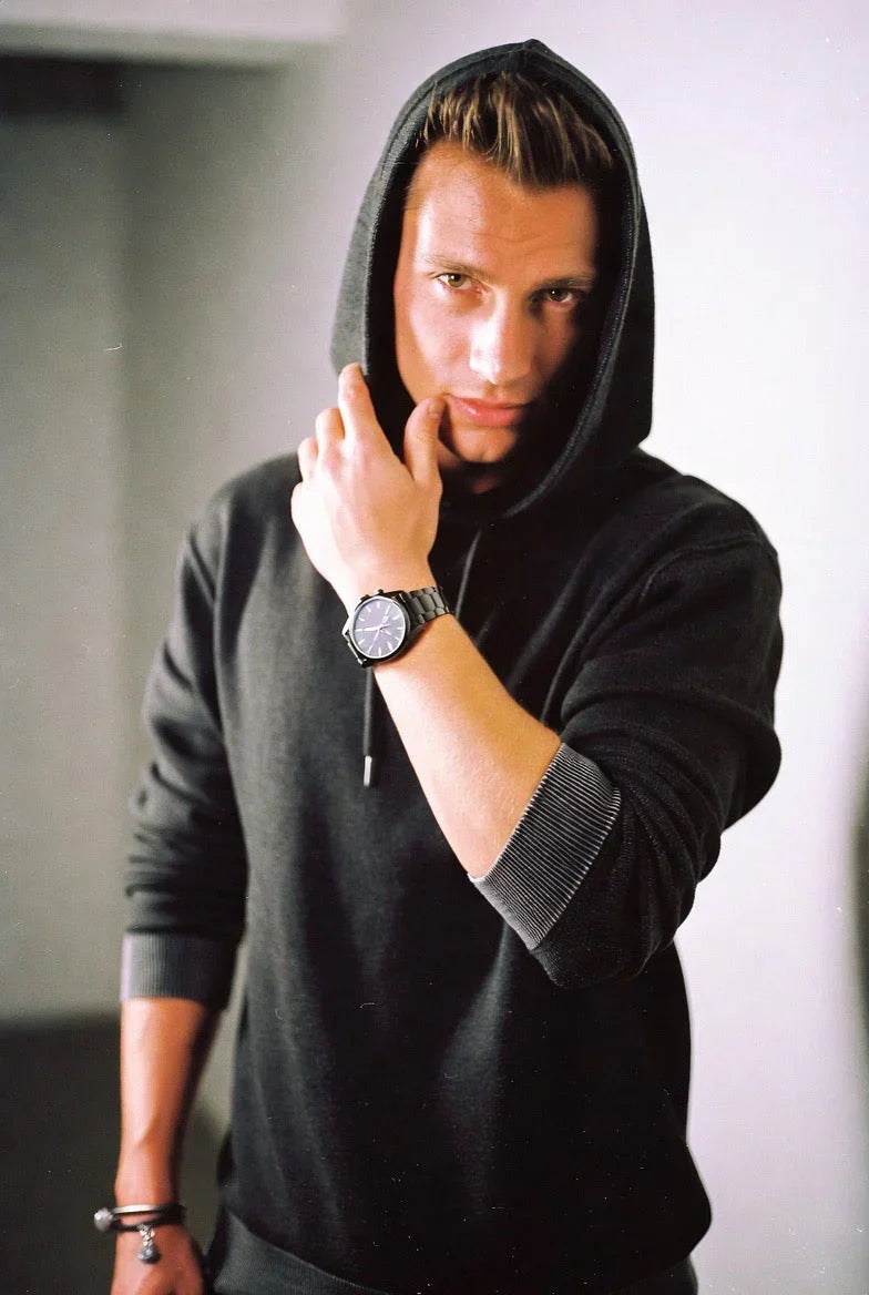 man in black hoodie wearing stylish black wristwatch and bracelets posing indoors