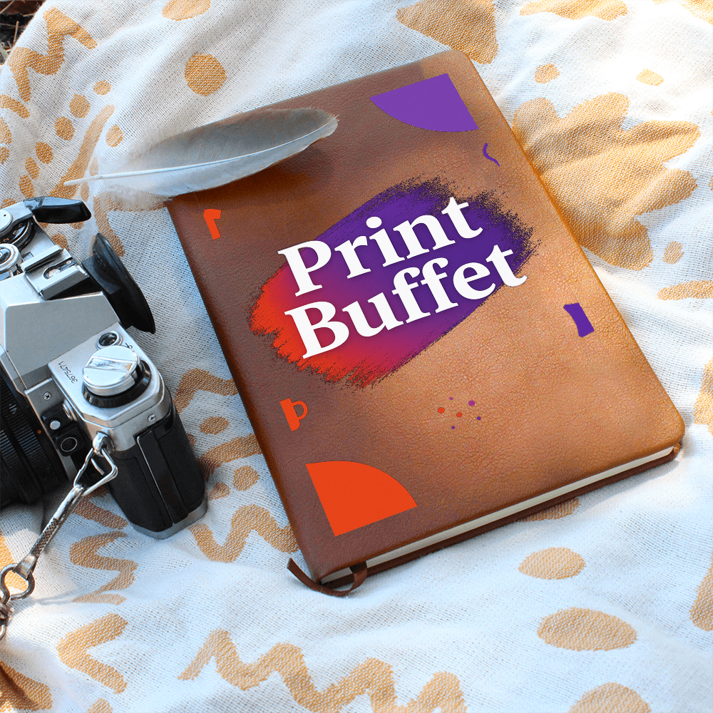 Vintage camera and feather next to brown leather notebook with Print Buffet text on patterned fabric