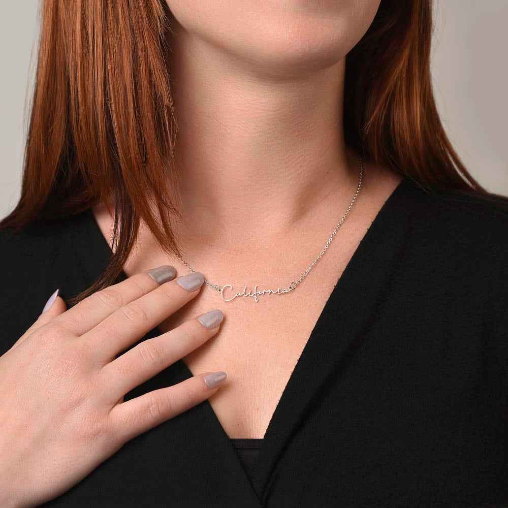 Close-up of woman with red hair wearing silver California name necklace and black top, hand touching necklace