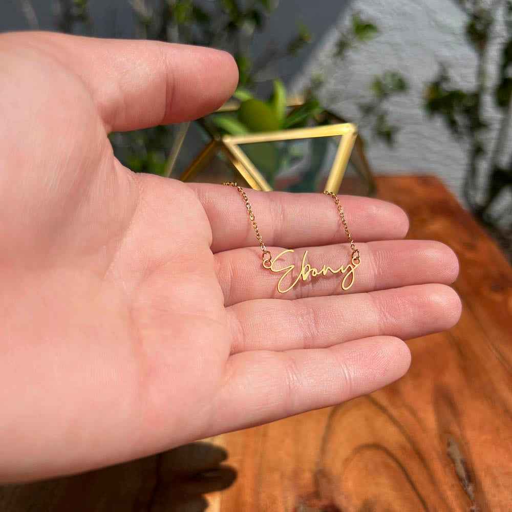 Hand holding a delicate gold name necklace with the word Ebony on a wooden surface