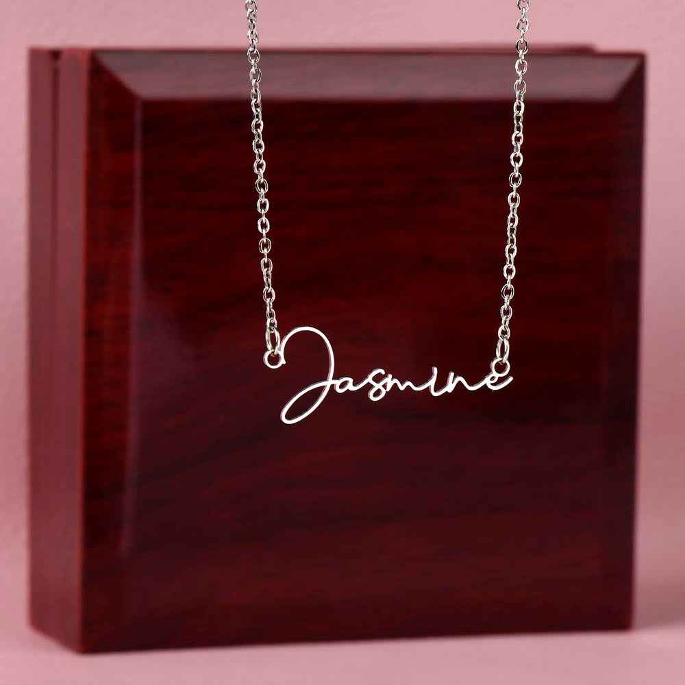Silver name necklace with cursive 'Jasmine' pendant displayed on dark wood box and pink background
