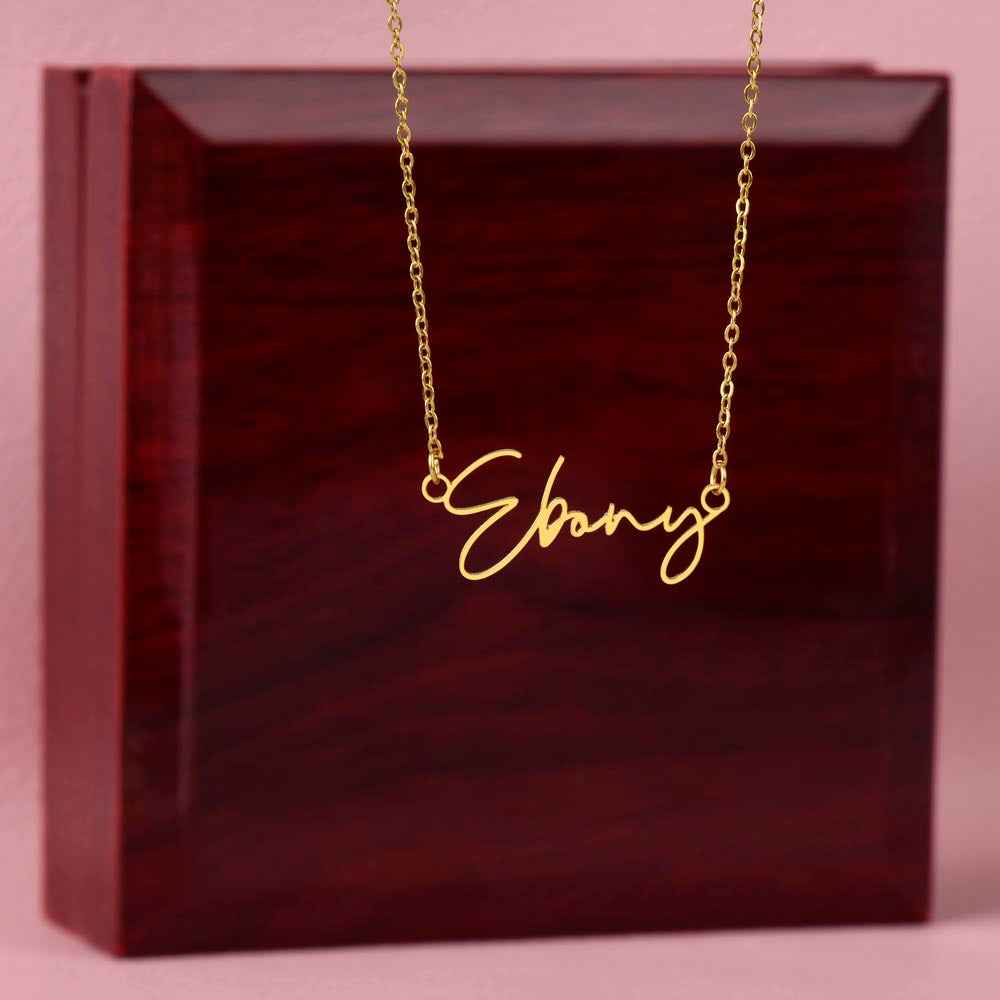 Gold name necklace with 'Ebony' pendant on chain against dark wooden block and pink background