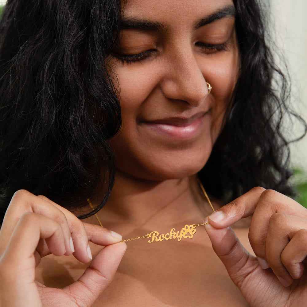 Smiling woman holding a gold necklace with the name 'Rocky' and a paw print pendant
