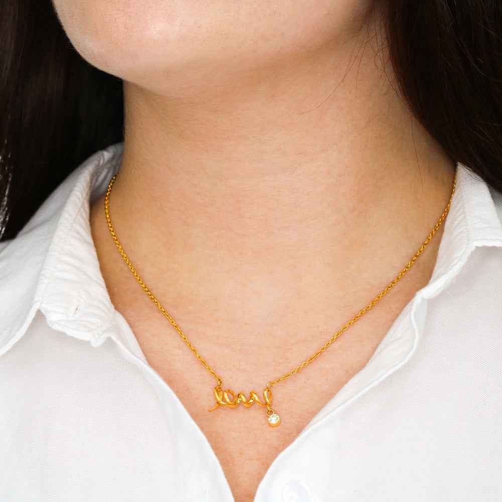 Close-up of gold love script necklace with small diamond pendant on woman wearing white shirt
