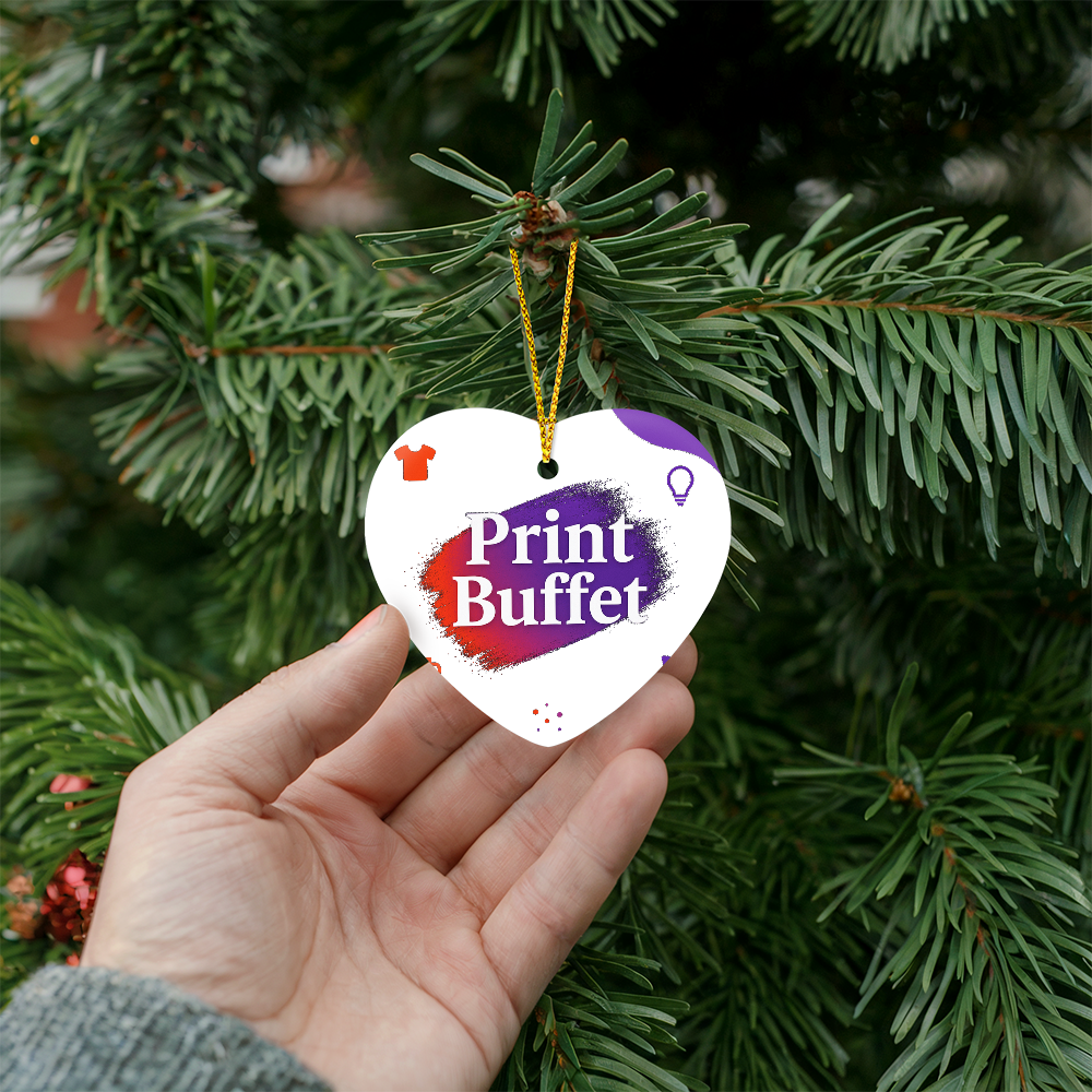 Hand holding heart-shaped Christmas ornament with Print Buffet logo on green pine tree